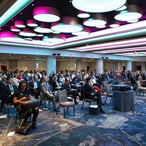 Large conference room with seated attendees spaced apart, modern ceiling lights with pink and blue accents, and a carpeted floor.