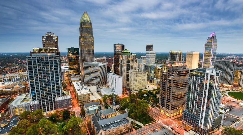 Aerial view of Charlotte, North Carolina downtown skyline at dusk with illuminated modern skyscrapers, streets, and green spaces under a partly cloudy sky.