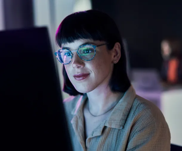 Young woman with glasses and short dark hair working on a computer in a dimly lit room with a soft glow on her face.