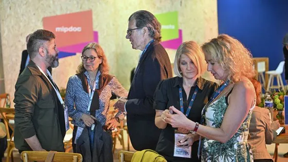 Five people engaged in conversation at a MIPDoc event, standing indoors with colorful event banners blurred in background, some wearing event badges.