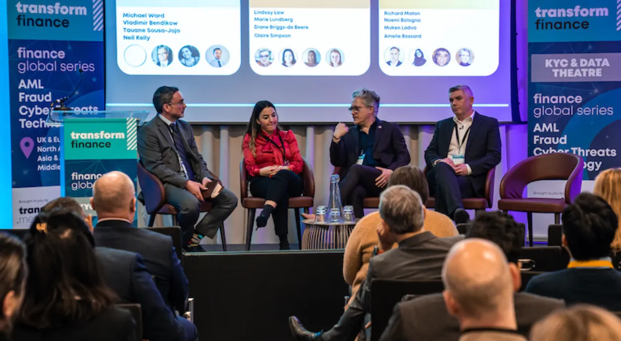 Four panelists seated on stage participating in a discussion at a Transform Finance event, with a screen behind them displaying names and photos of speakers and attendees watching in the foreground.