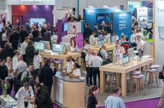Busy exhibition hall with many people interacting at booths and information desks, several computer screens on tables, and large blue and white promotional displays in the background.