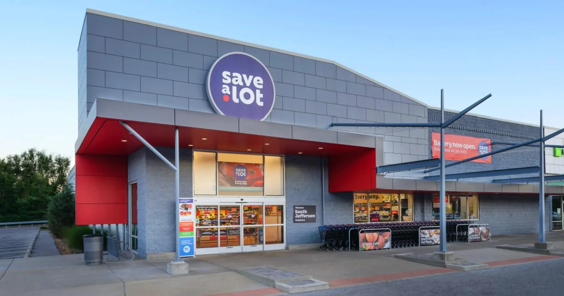 Exterior of a Save-A-Lot grocery store with a large purple and white Save-A-Lot logo above the entrance, red accent panels, shopping carts lined up on the right, and clear skies in the background.