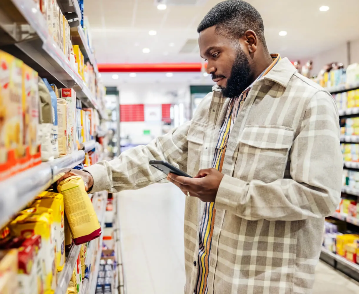 Man with a beard in a checkered jacket looks at a packaged product on a grocery store shelf while holding a smartphone in his other hand.