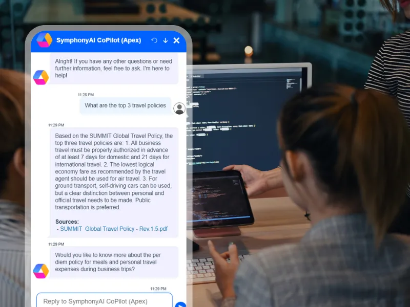 Chat window with SymphonyAI CoPilot (Apex) overlaying a photo of two people working at a desk with a computer and tablet showing code, discussing top three business travel policies including authorization times, economy fares, and ground transport preferences.