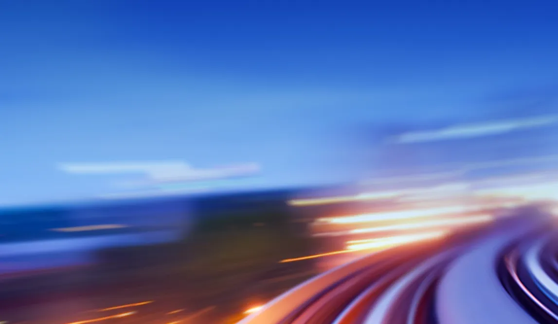 Blurred dynamic light trails on a curved road or highway at dusk, suggesting motion and speed under a clear blue sky.