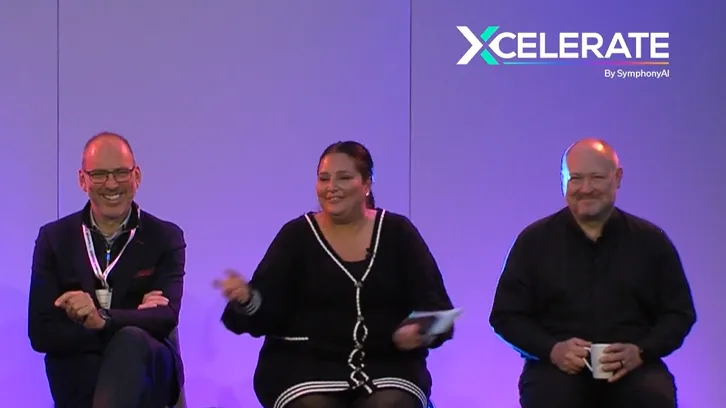 Three people seated on stage against a purple background at an Xcelerate by SymphonyAI event, with the logo in the top right corner; the person on the left is smiling with arms crossed, the person in the center is gesturing with one hand and holding papers, and the person on the right is holding a cup and smiling.