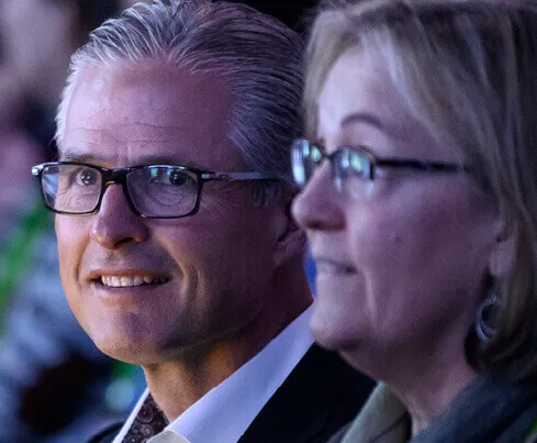 Close-up of two middle-aged people wearing glasses at an event; a man with gray hair and a woman with short blonde hair are seated next to each other.