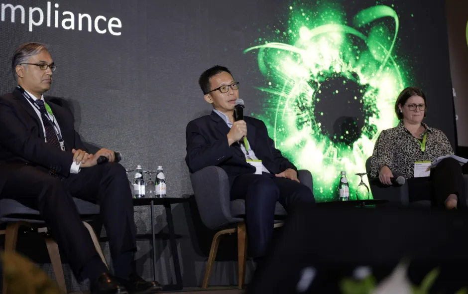 Three people seated on stage participating in a panel discussion at a conference, with one man in the center speaking into a microphone, a man on the left and a woman on the right holding microphones, against a backdrop featuring the word 'compliance' and a green abstract graphic.