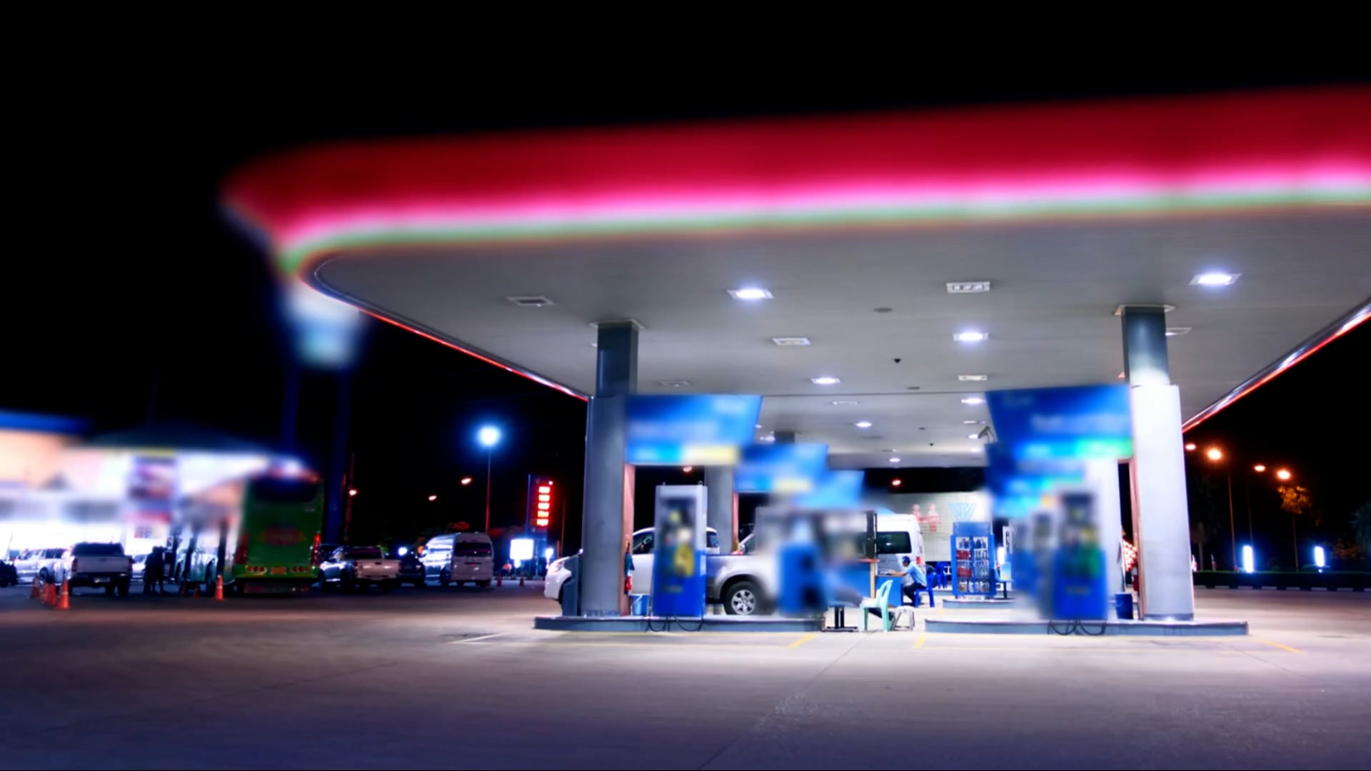 Nighttime view of a gas station with illuminated canopy and fuel pumps, several vehicles parked, and people visible near the pumps and a seating area.