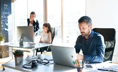 A man working on a laptop at a desk in a bright office while two women work together at a computer in the background near large windows.