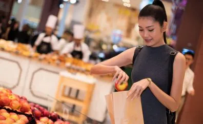 A woman placing an apple into a paper bag while shopping in a grocery store with fruit displays and staff in the background.