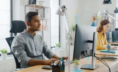 Man focused on work while typing on a computer keyboard at a desk in a bright office with a woman working in the background.