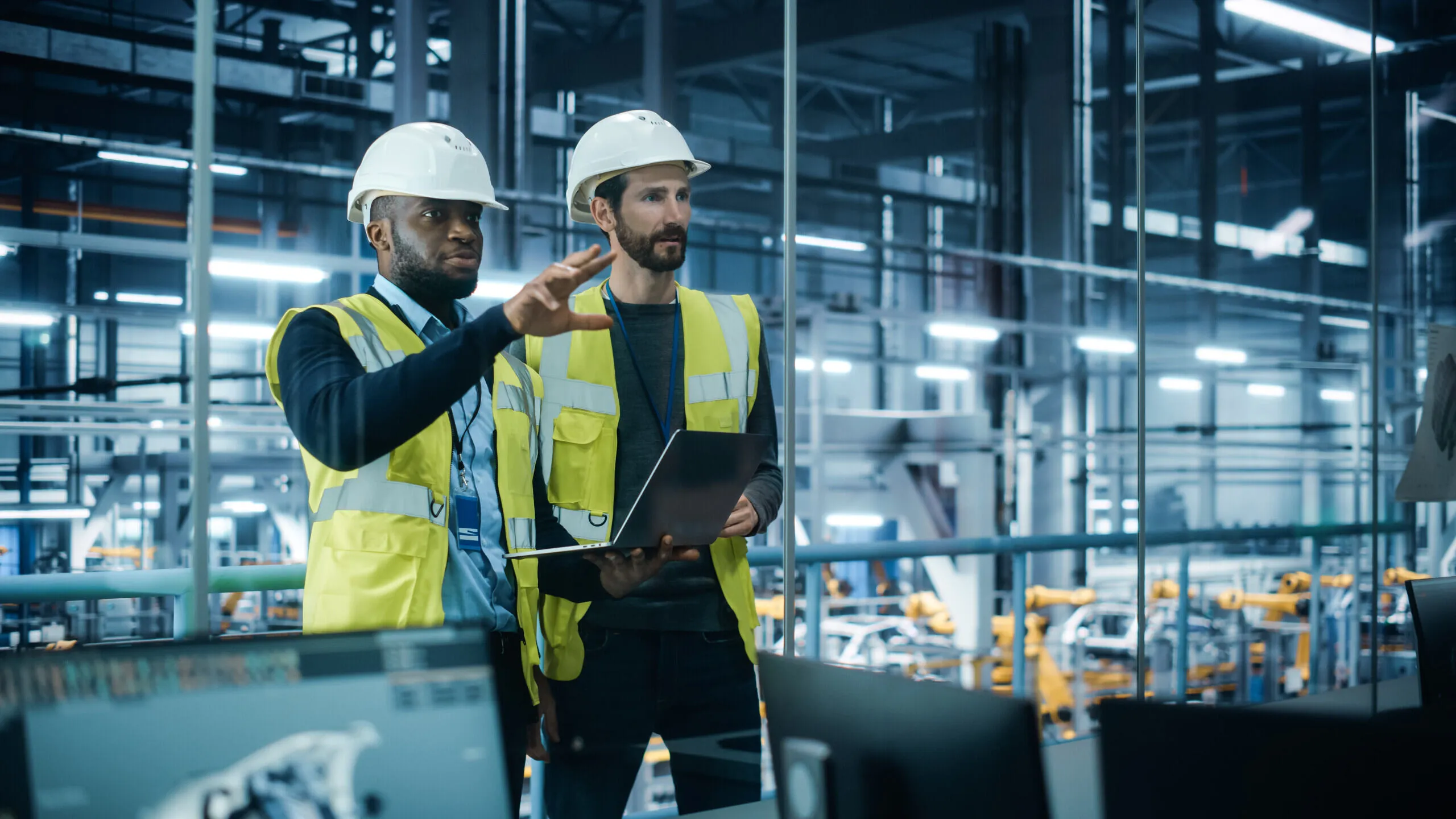 Two male engineers wearing white hard hats and yellow safety vests stand inside a modern factory with industrial robots in the background; one engineer is holding a laptop while the other gestures with his hand.
