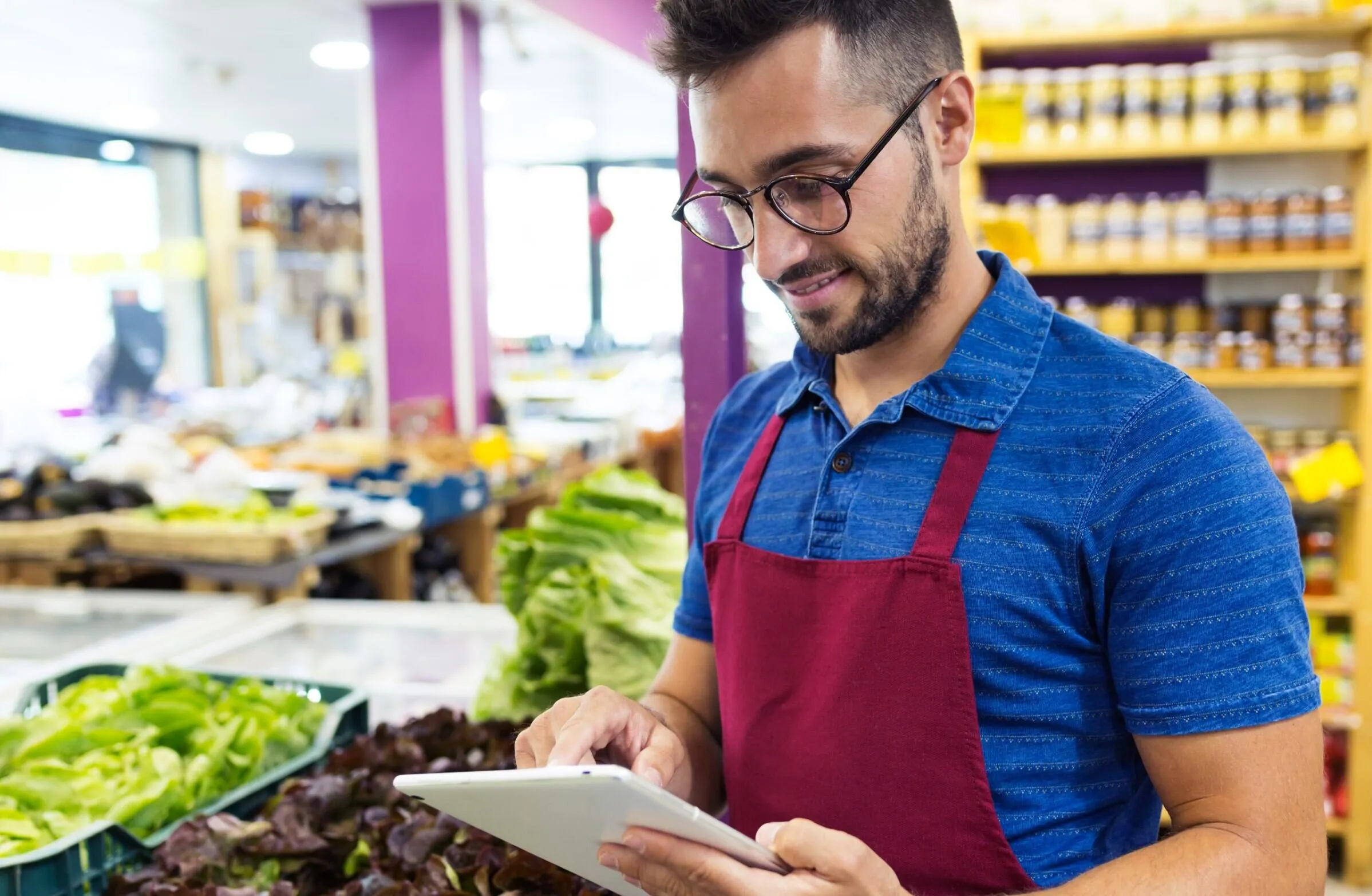 Man wearing glasses and a red apron standing in a grocery store produce section, using a tablet with fresh greens around him.