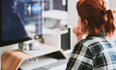 A person with red hair in a plaid shirt sits at a desk looking at a computer screen displaying a blurred figure.