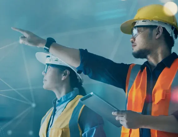 Two industrial workers wearing safety vests, helmets, and glasses; one worker points forward while the other holds a tablet, against a blue background with network-like light lines.