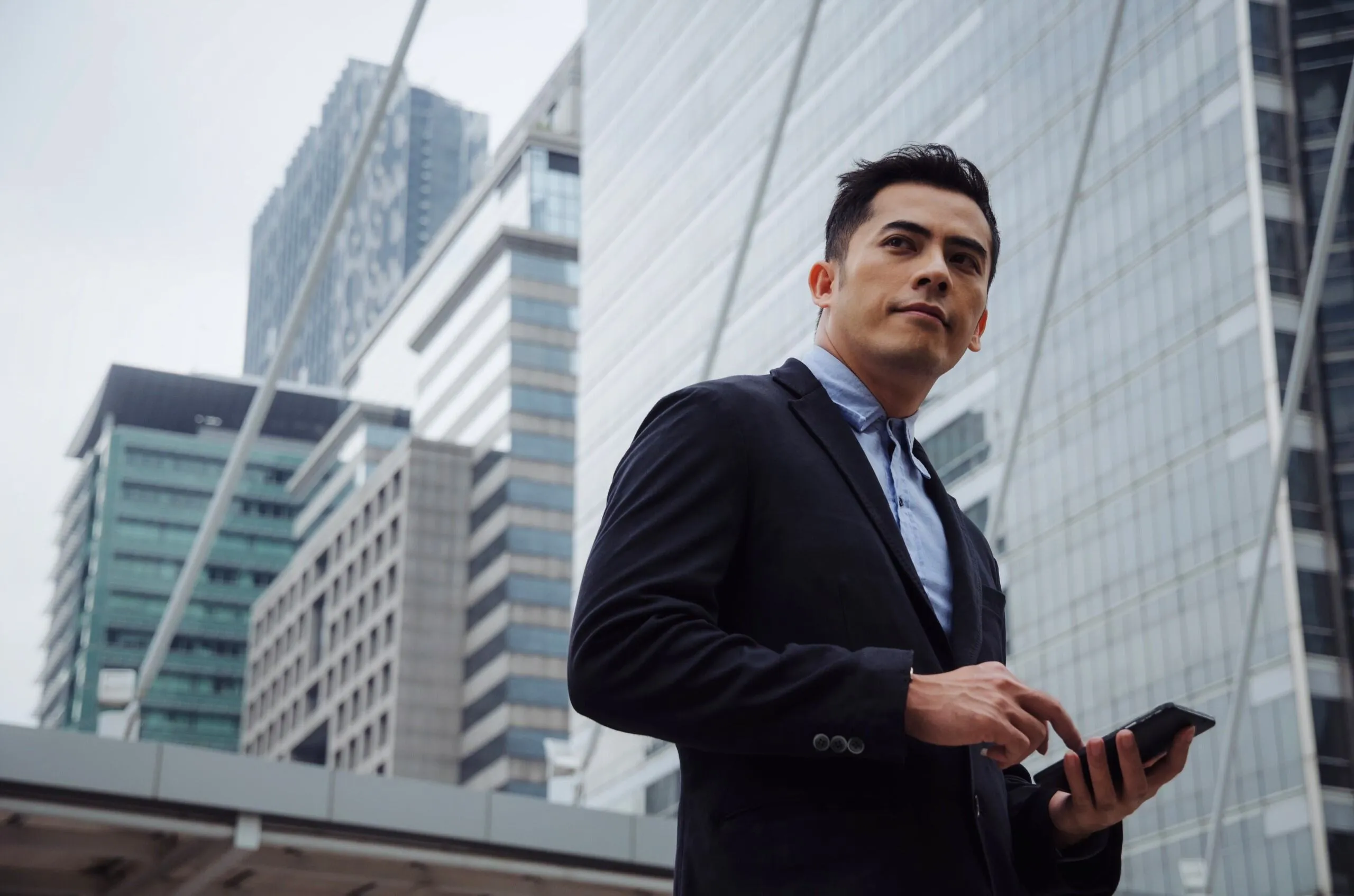 Young businessman in a black blazer and light blue shirt standing outdoors in a city with modern glass and concrete buildings in the background, holding and using a smartphone while looking to the side.