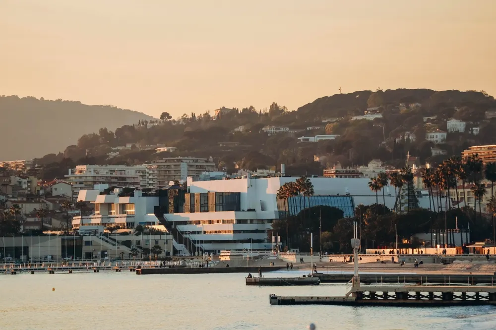 View of a coastal cityscape with modern white buildings, palm trees, and a mountainous background during sunset, with calm water and piers in the foreground.