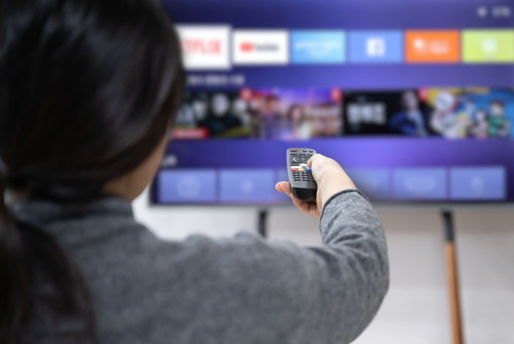 Person holding a remote control pointed at a smart TV displaying a streaming service interface with icons for Netflix, YouTube, and other apps.