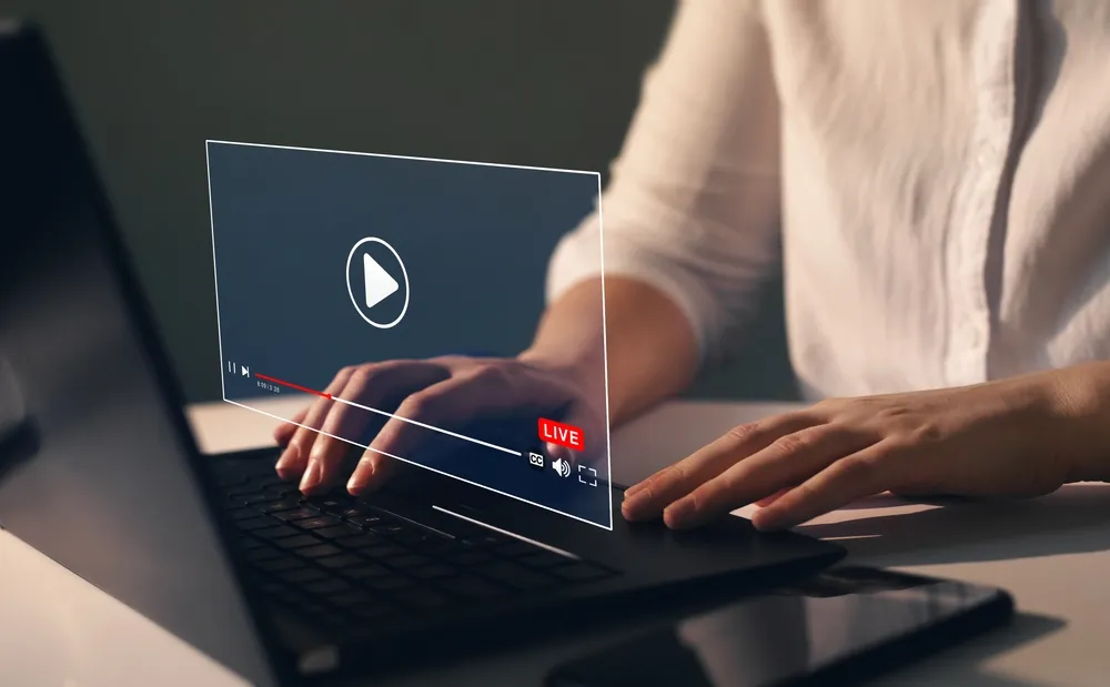 Person using a laptop with a transparent interface of a video player showing a live streaming indicator and playback controls floating above the keyboard.