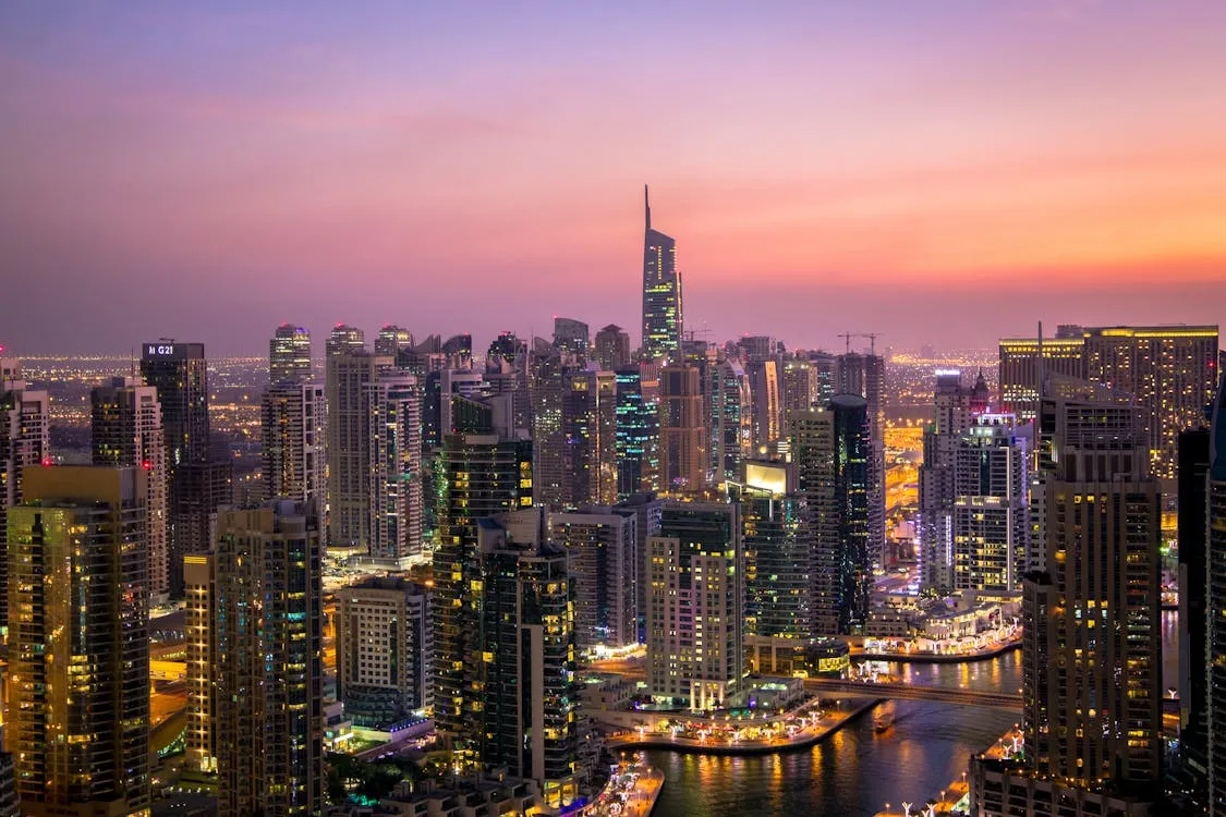 Dubai city skyline at dusk showing numerous illuminated modern skyscrapers surrounding a winding waterway, with a colorful purple to orange gradient sky above.