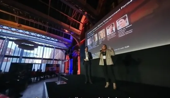 Two presenters stand on a stage in front of a large screen displaying speaker details at an indoor event with high windows and visible audience seats.