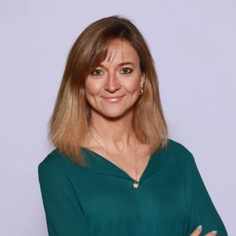 Smiling woman with shoulder-length light brown hair wearing a teal blouse and a heart-shaped pendant necklace against a plain light background.