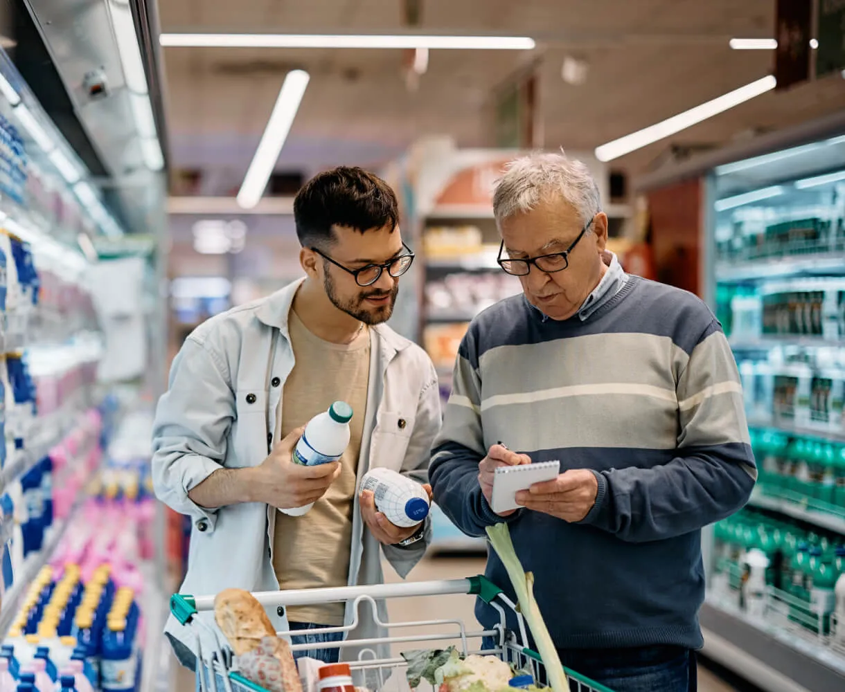 A younger man holding two bottles, comparing their labels, while an older man writes on a notepad in a grocery store aisle with shelves of products and a shopping cart with vegetables and bread in front of them.
