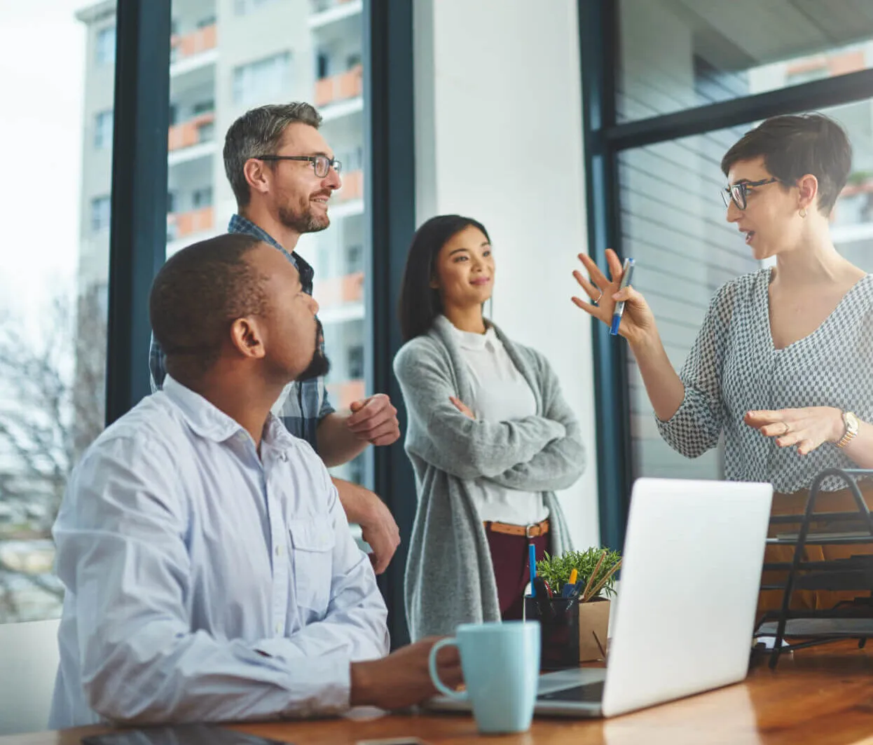 Four diverse colleagues having a discussion in a modern office, with one woman standing and explaining something using hand gestures while the others listen attentively near a laptop and coffee mug on the table.