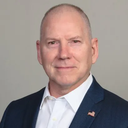 Portrait of a middle-aged man with a bald head wearing a dark suit jacket, white shirt, and a small American flag pin on his lapel, against a plain gray background.