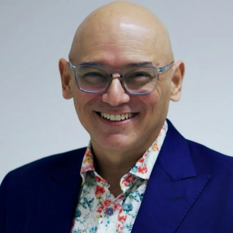 Smiling bald man wearing clear-framed glasses, a purple blazer, and a white shirt with a colorful floral pattern, against a plain light background.