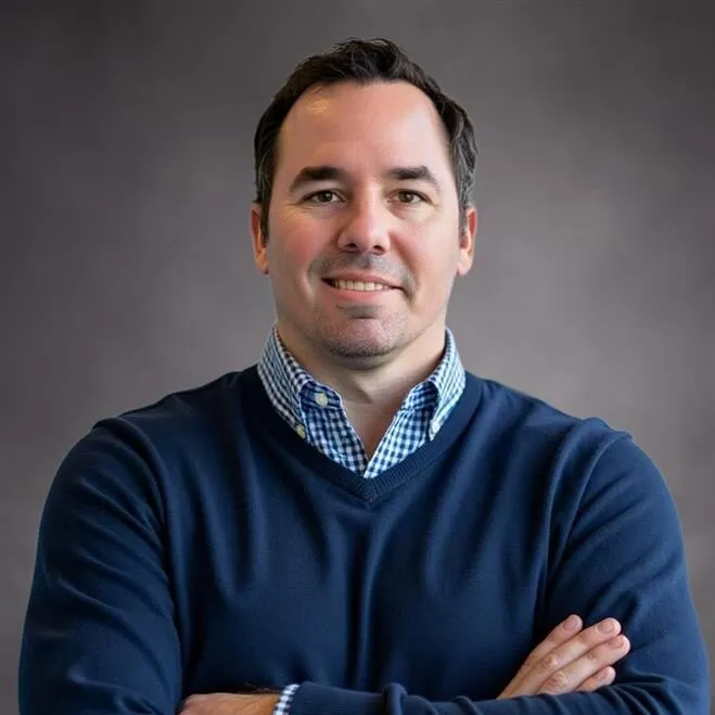 Portrait of a man with short dark hair wearing a blue sweater over a blue and white checkered shirt, smiling with arms crossed against a plain gray background.