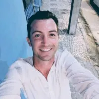 Smiling man with dark hair and light skin wearing a white shirt taking a selfie on a stone-paved street at night.