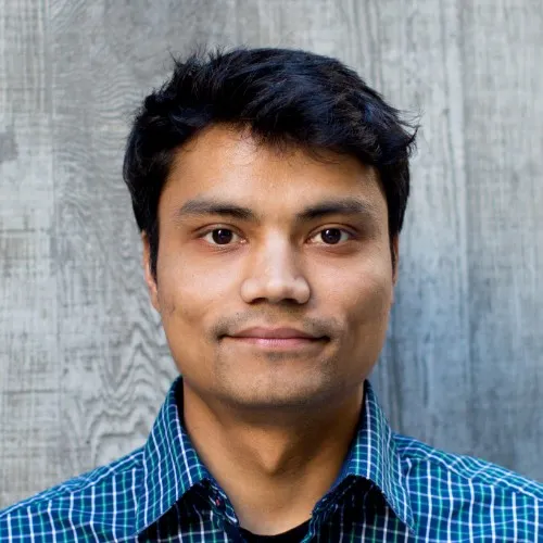 Young man with short black hair wearing a blue and green checkered shirt, standing against a textured gray wooden background.