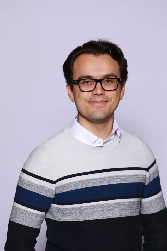 Smiling man with dark hair and glasses wearing a white shirt under a gray, blue, and black striped sweater against a plain light background.