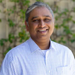 Smiling middle-aged man with gray hair wearing a white and blue checkered shirt standing outdoors with a blurred leafy background.