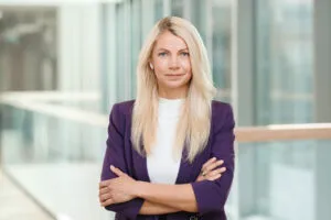 Confident blonde woman with long hair in a purple blazer and white top standing with arms crossed in a modern office building with glass walls.