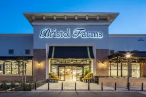 Exterior of a Bristol Farms store at dusk with illuminated sign above the entrance, large windows, and decorative lighting along the facade.