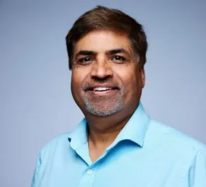 Portrait of a middle-aged man with brown hair and a trimmed beard, wearing a light blue collared shirt, smiling against a plain gray background.