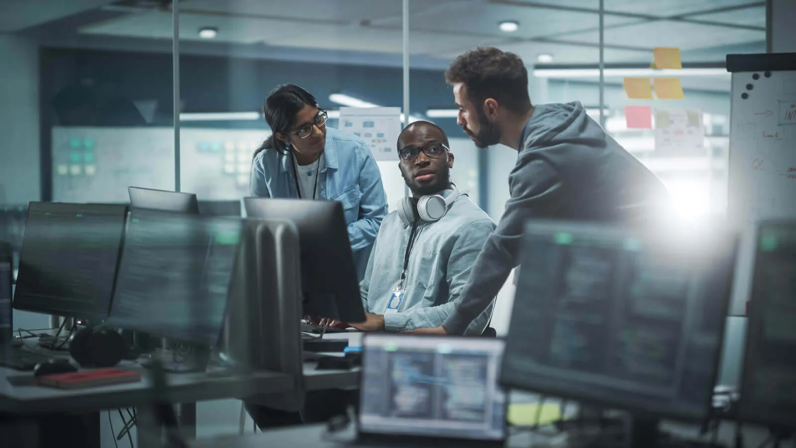 Three diverse professionals collaborating in a modern office with multiple computer monitors, one seated man with headphones around his neck listening while two colleagues, a woman and a man, discuss work near his desk.