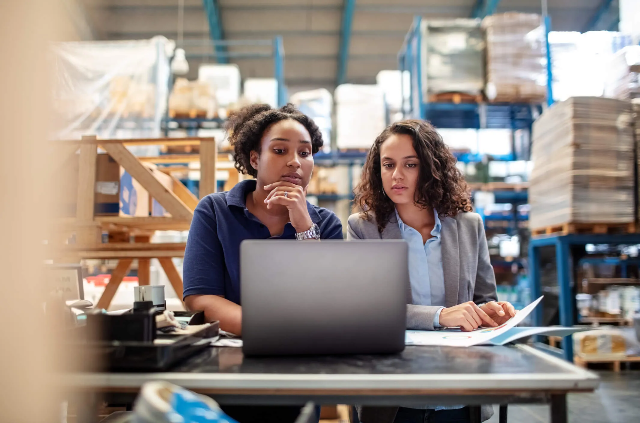 Two women working together in a warehouse, looking at a laptop on a table with shelves and boxes in the background.