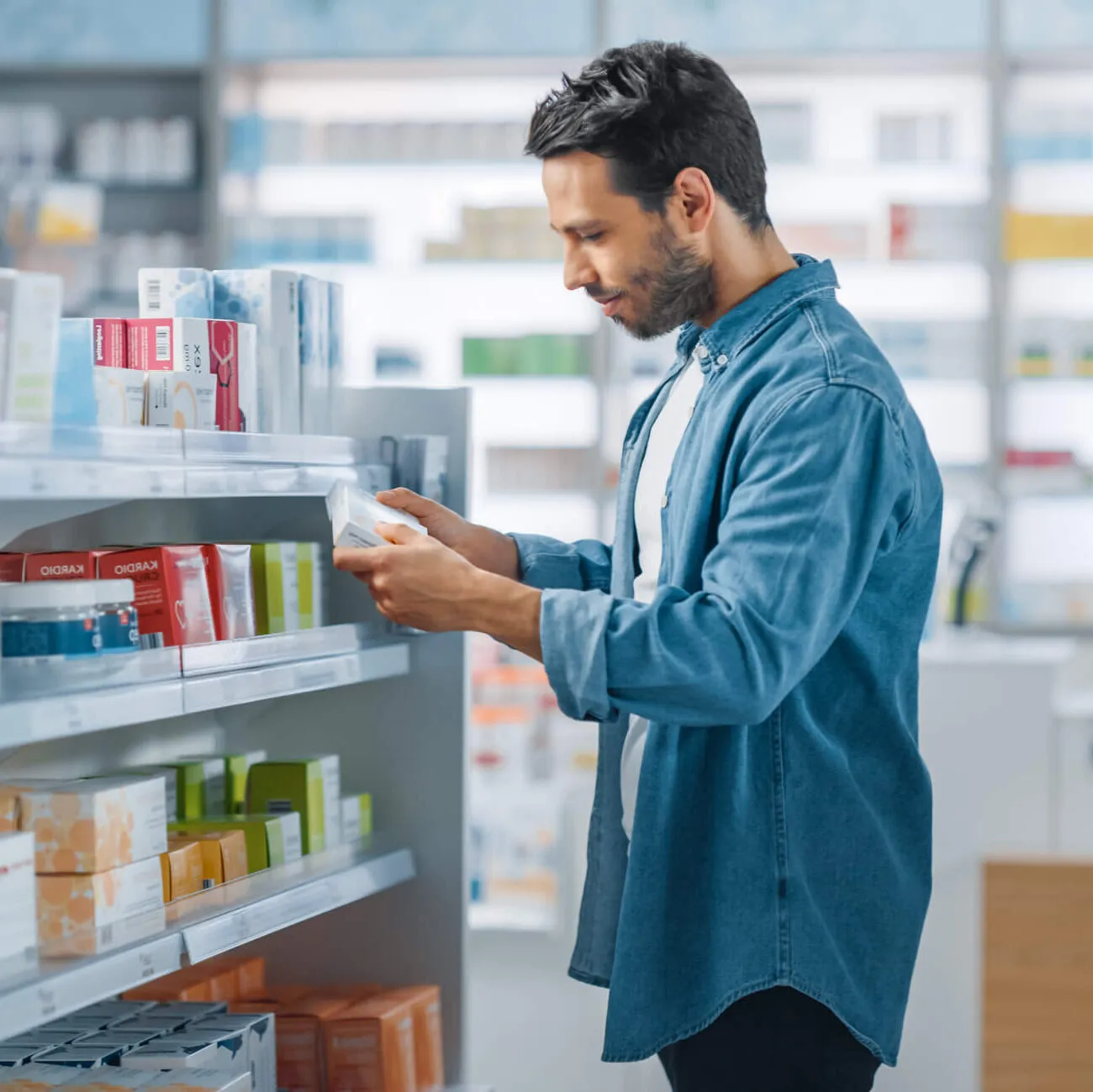Man in a denim shirt reading the label of a product in a brightly lit pharmacy with shelves of medications.