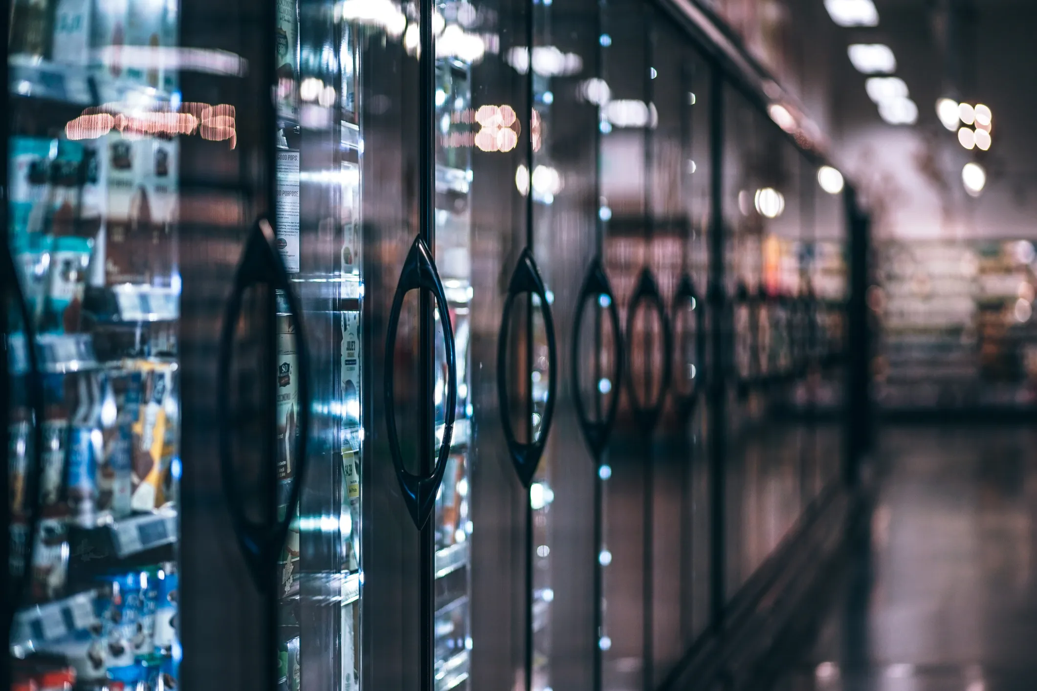 Row of glass-door freezers in a grocery store with various frozen goods visible inside under bright overhead lighting.