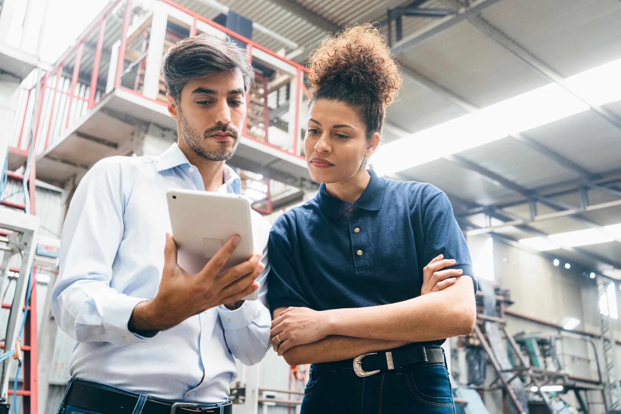 A man in a light blue shirt and a woman in a dark blue polo shirt are standing in an industrial warehouse, looking intently at a tablet the man is holding.