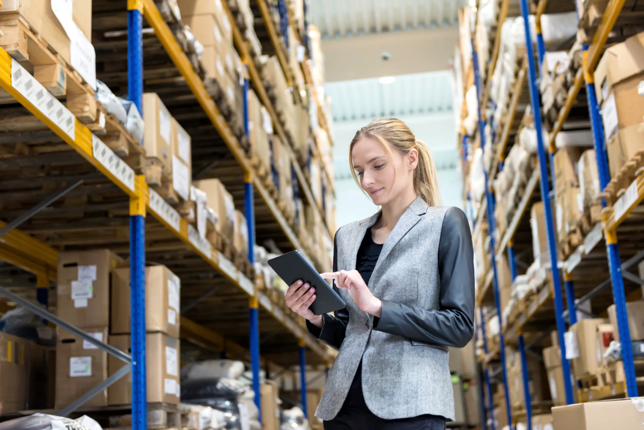 A woman in a gray blazer with black sleeves using a tablet in a warehouse aisle with tall shelves stocked with boxes and packages.
