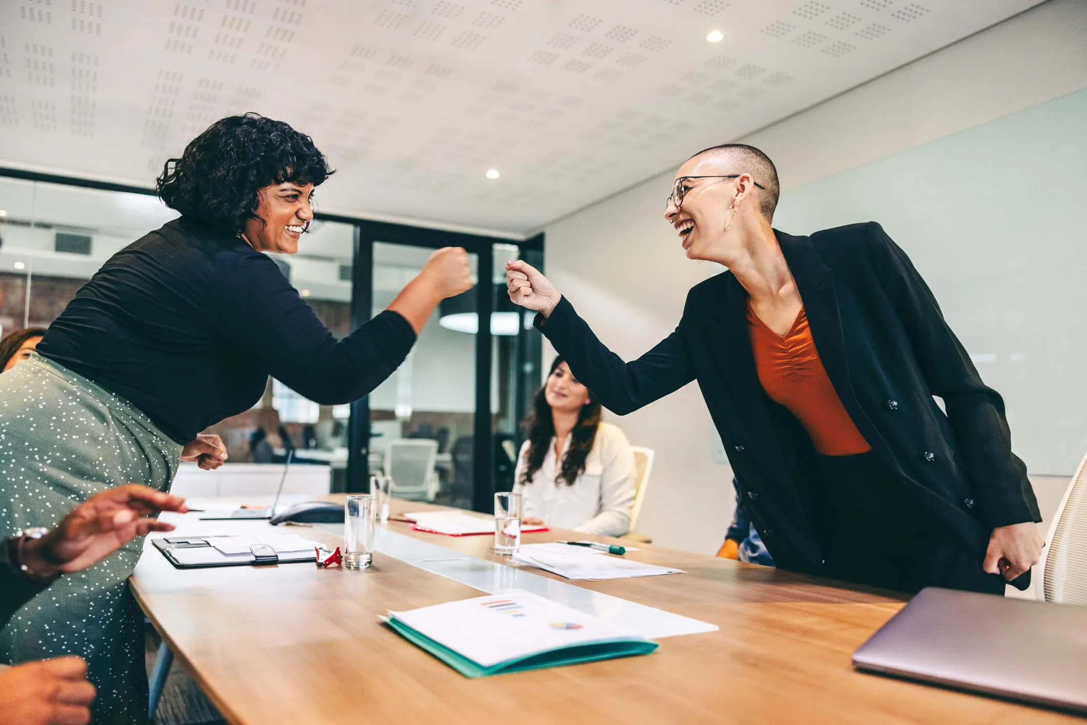 Two women in a modern office conference room smiling and giving each other a fist bump across a wooden table with documents, glasses of water, and a laptop.