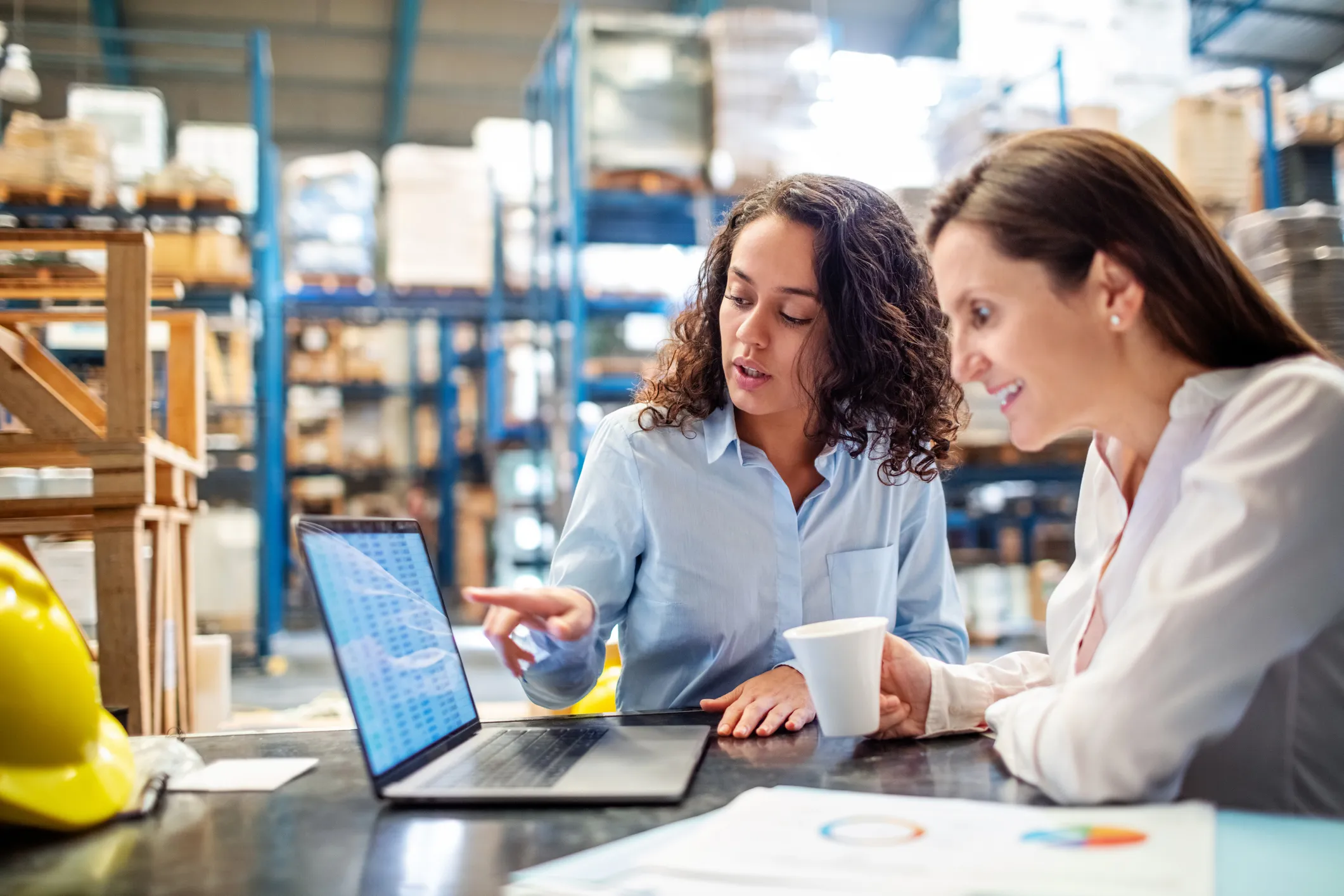 Two women in a warehouse discuss information shown on a laptop screen while one points at the laptop and the other holds a white cup, with industrial shelving and a yellow safety helmet in the background.