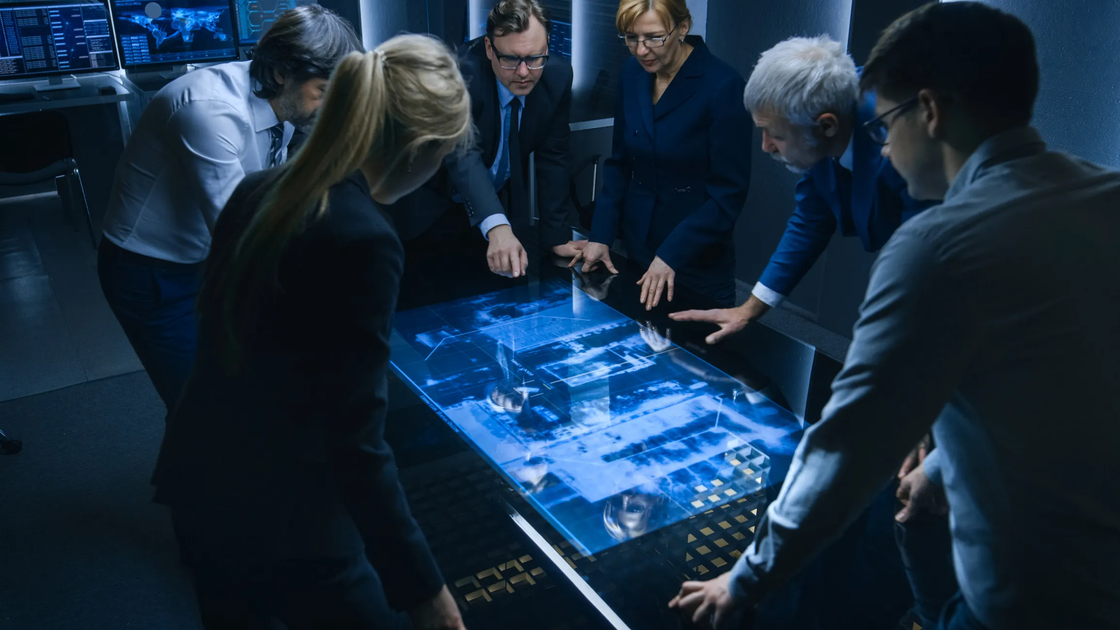 A team of six professionals in business attire gathered around a large interactive digital table displaying blue-toned data and maps, collaborating in a high-tech control room with multiple monitors in the background.