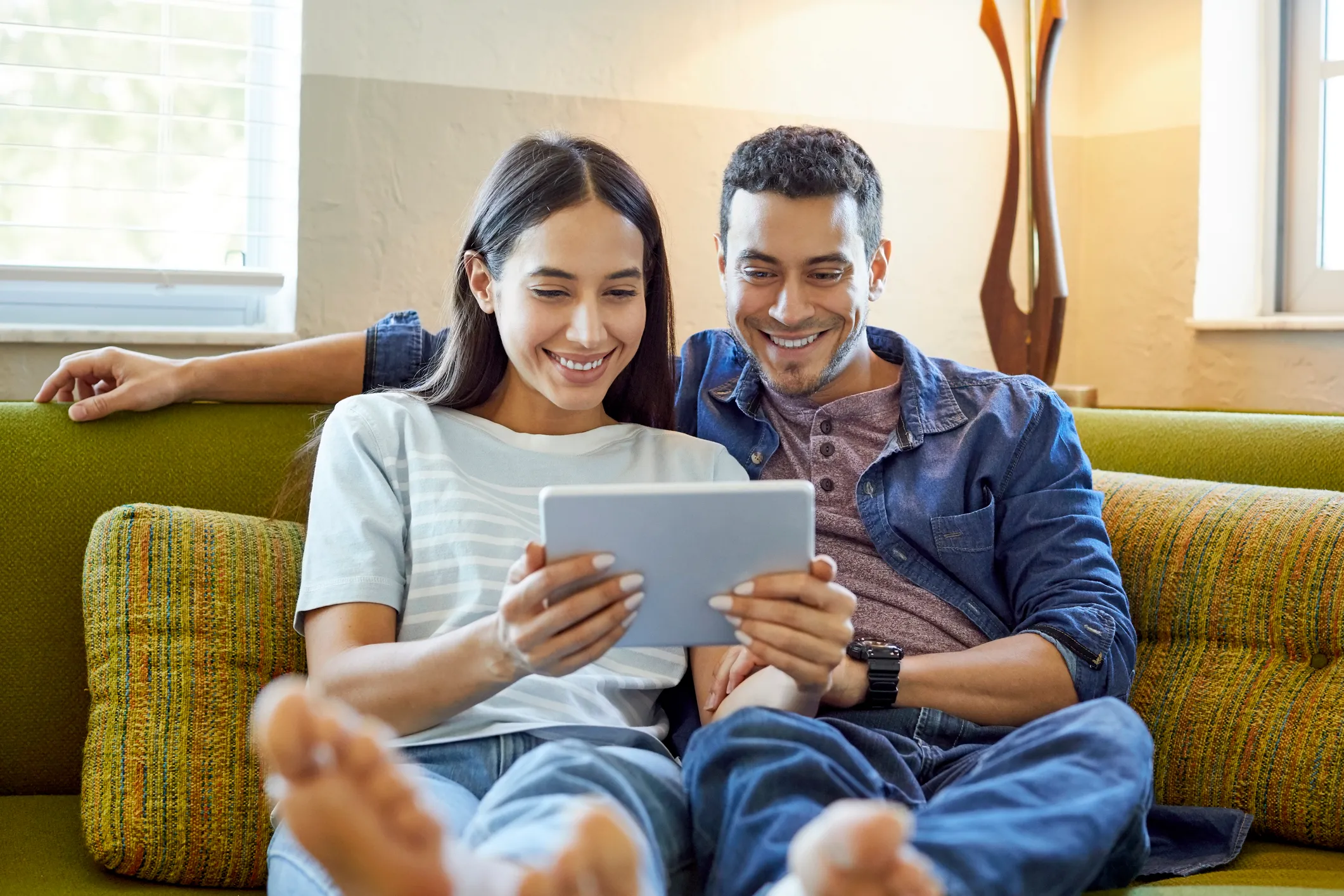 Smiling couple sitting barefoot on a green couch watching a tablet together in a bright room with windows and wooden decor.